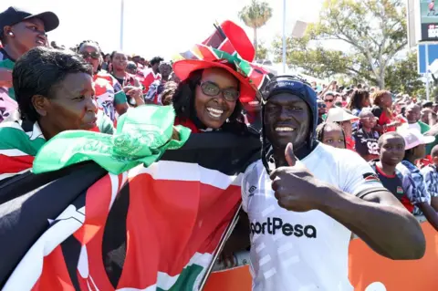 PAUL KANE / GETTY IMAGES A man poses with his thumbs up. Next to him a woman waves the Kenyan flag.