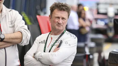 Allan McNish standing with his arms folded. He has light brown spiky hair and a look of concentration on his face. He is wearing an F1 lanyard and a white Audi Revolut long-sleeved top.