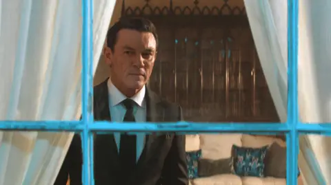 WORLDPRODUCTIONS/PA Evans is staring past the camera through a window with a chipped white frame. He wears a black suit and tie, with a white shirt.