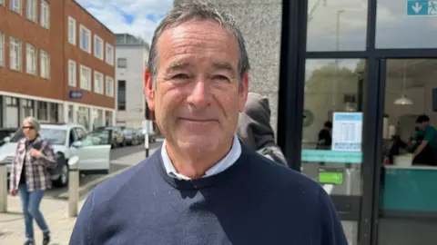 A man stood wearing a navy jumper and light blue shirt stood in front of a shop. 
