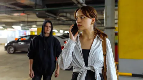 A stock image of a woman wearing a black vest underneath a striped shirt, which is tied at the waist. She has brown hair in a ponytail and is carrying a brown bag on her shoulder. She is walking through an underground car park while a man in a black hoodie follows her at a close distance.