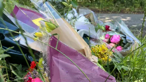 Stuart Woodward/BBC Bunches of flowers are lying on the green grass. There are several bouquets, showing yellow, red and pink petals.
