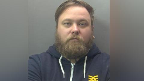 Grant Brownlow in police custody wearing a navy blue zip-up hoodie. He has a beard.