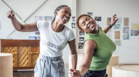 Marc Brenner Two women in a rehearsal space. The one on the left has black hair pulled back tightly behind her head and wears a white T-shirt with black writing on it, only part of which can be made out. On the right is a woman with short brown hair and wearing a sleeveless green top and black trousers. Behind them is the back of a wooden piano and several photographs on the wall.

