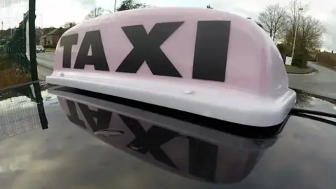 A close up generic image of a white taxi light with black writing which says TAXI on top of the roof a black car