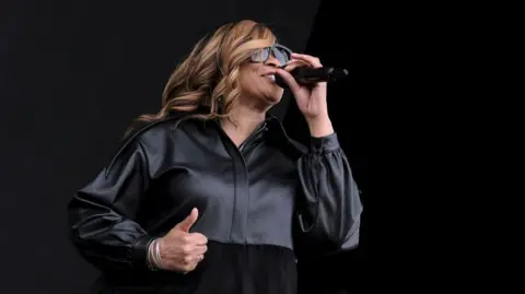 Getty Images Gabrielle singing into a microphone. She has long blonde hair, wearing black sunglasses and a black outfit.