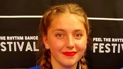 Family photo Chloe Longster smiles for the camera. She wears her hair in pigtails with stud earrings and red lipstick. The picture is taken at a dance festival where he was awarded a certificate.