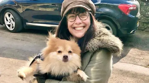 Lisa Burtenshaw Lisa Burtenshaw is pictured holding a fluffy cream-coloured dog. Both of them are looking directly at the camera and Lisa is smiling widely. She is standing on a pavement and there is a black parked car behind her on the road. Lisa is wearing a brown suede peaked cap and a khaki coat with a fake fur collar.
