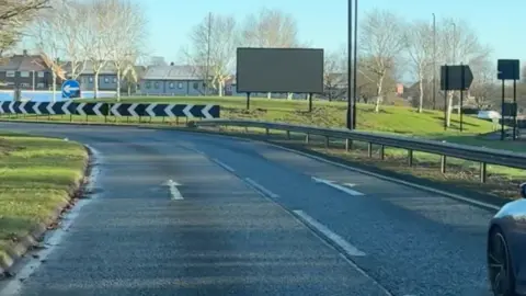 A road with a grass verge to the left leads to a roundabout where a large black LED display has been installed.