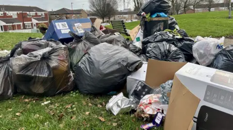 Una pila de cajas y basura apiladas unas sobre otras en un parque al lado de un banco.