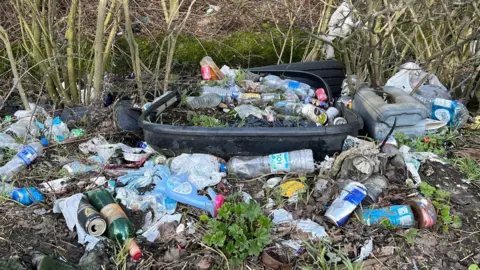 A wooded area where the muddy ground is covered in litter, including glass and plastic bottles, plastic bags, cans, tins and what appears to be a black plastic bin, which is partially buried and also covered with litter. Bare shrubs are in the background.