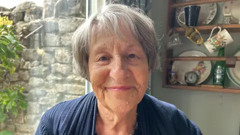 A woman with short grey hair, wearing a blue cardigan, sitting in her kitchen and smiling at the camera