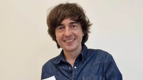 Danny Robins smiling wearing a denim shirt. It is a head and shoulders shot.