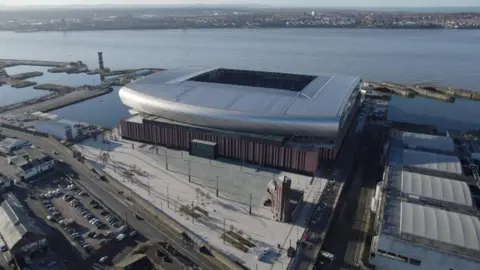 An aerial view of Everton Football Club's new Bramley-Moore Dock stadium. It is a large silver structure with a curved roof. The stadium is on the banks of the River Mersey.