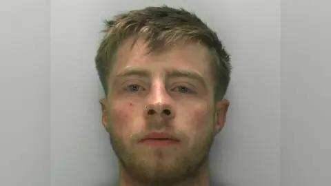 A mugshot of a man in his early 20s. He has shorter dirty blond hair and blond facial hair. He has a nose ring on one nostril. He has a blank expression and is standing in front of a plain grey background.