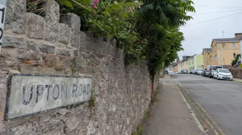 A close up image of a road sign that is attached to a stone wall. It shows the road and a pavement with cars and houses in the background. 