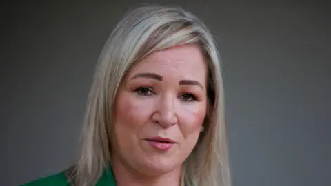 PA Michelle O'Neill is wearing a green suit. She's talking to the media. She has shoulder length blonde hair and is wearing pearl earrings.