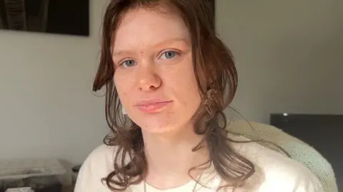 A person with shoulder-length brown hair wearing a white t-shirt looks at the camera while sitting in a room with white walls and a television. 