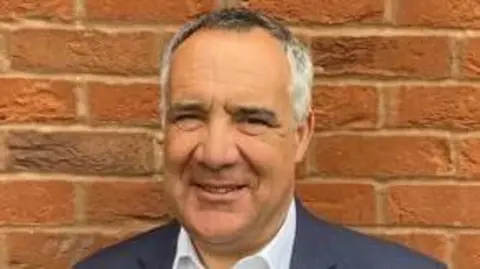 Herefordshire Council A clean-shaven man with partially receding hairline, which is dark grey and white. He is wearing a white shirt and a navy jacket while stood in front of a red brick wall.