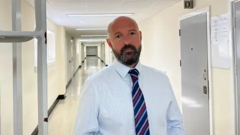 A bald man with a short dark beard look at the camera. He is wearing a light blue shirt with a dark blue tie with pink, red and a lighter blue stripes. He is in the corridor of a prison.