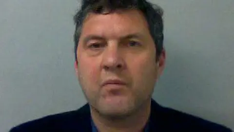 Neil Stafford mugshot. He has short dark hair, and is wearing a black blazer over a blue shirt.