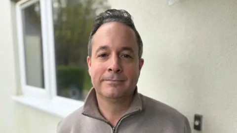 Nick West stands in front of his home next to the doorbell. He is a middle aged man with grey hair. He is wearing a grey jumper. 