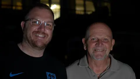 Jamie Niblock/BBC Andy Gore, who is wearing a navy England top, and dad Clive, who is wearing a green polo top. Both are smiling at night.