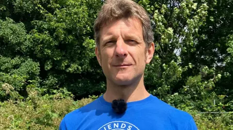 Ian stands for a photo. He is wearing a blue top. He has short brown hair. Taken on a sunny day.