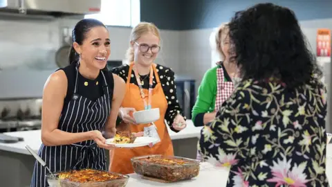 Getty Images Duchess of Sussex smiles as she presents a plate of food to another woman in a floral-patterned shirt. Two other woman grin next to her.