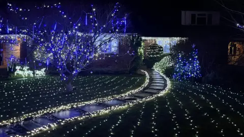 Adrian Harms/BBC Another house in Milden Gardens, Frimley Green, covered in Christmas decorations. This house has a series of white lights trailed across the front garden, and blue lights in a tree in the garden.