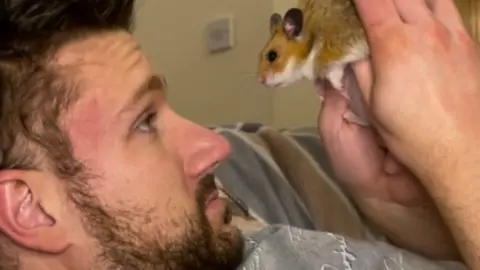 Chris is lying on the sofa holding Popcorn the hamster in the air. Popcorn is looking straight at him.