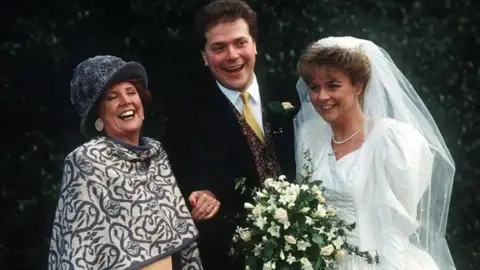 ITV/Rex Features Cilla Black wearing a grey hat and grey and cream shawl, stood next to Alex Tatham wearing a black suit, white shirt and yellow tie, stood next to Sue Tatham dressed in a wedding veil and dress holding a bouquet of white flowers on their wedding day. All of them are smiling and looking happy.