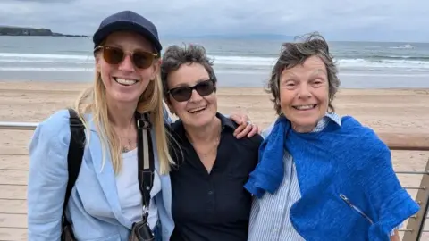 Helen Quinn is wearing a blue striped shirt and navy blue shawl.
She has short brown hair and is standing near her two friends who are both wearing sunglasses.