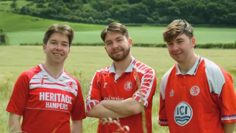 Tom Banks and Paul Thompson Theo, Oscar and Isaac are stood in the middle of a field with a forest behind them in the distance. They are all smiling and wearing red and white Middlesbrough shirts from different eras.
