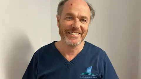 Vikki Irwin/BBC Dr Morton is wearing blue medical scrubs with a Beccles medical centre written on the left side. He is smiling at the camera and has a beard. He is photographed against a white wall 