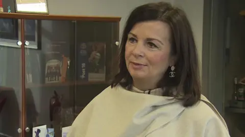 Christine Fearon, wearing a cream poncho/shawl and chanel diamond earrings. She has short brown hair and is standing in the school foyer with glass cabinets behind her