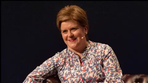 Nicola Sturgeon in a patterned blouse, smiling with a lip mike fitted