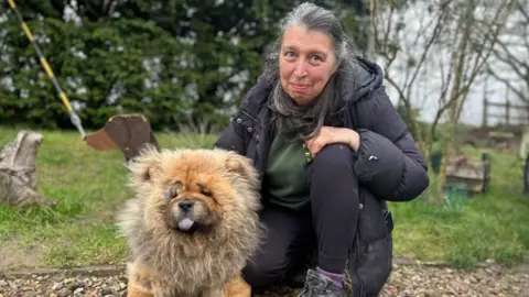 Julie Shelton, holding Cato the dog. Julie is crouching down, with long hair, wearing a black coat, trousers and green top. She is looking straight at the camera. Cato is a Chow Chow dog, and has his tongue sticking out. 