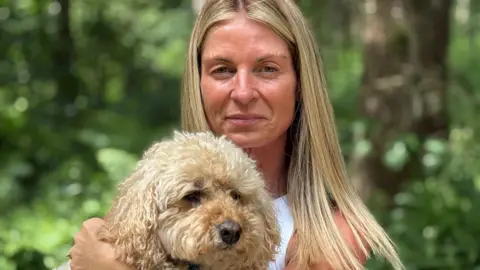 A blonde woman holds up her blonde Cavapoo dog.