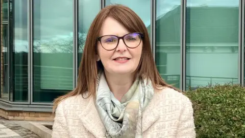 Dr Caoimhe Archibald stands in front of a glass fronted building at Ulster University in Derry, she has shoulder length brown hair, and is wearing glasses, a cream overcoat and scarf.