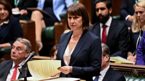 Jessica Taylor/Reuters Rachel Reeves standing at the dispatch box in the House of Commons. She is changing pages in her documents. Labour politicians can be seen in the benches behind her.