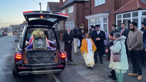 A black hearse with the boot open and a framed portrait of Manjula Sood propped up against her coffin. A crowd of people and undertakers are stood beside it.