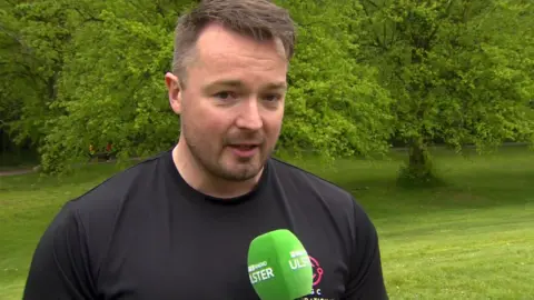 Graham Rennie, a man with short, fair hair, talks to a BBC TV crew in the grounds of Stormont. He is wearing a black crew-neck t-shirt and is talking into a green microphone. There is a lawned area covered in mature trees in the background.