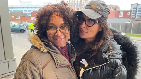 Vaneca Sinclair (left), a woman with short brown curly hair and glasses wearing a light brown coat with a fur hood, stands in a doorway facing the road next to Savita Kapur (right) with long brown hair, glasses, a dark green cap and a black puffer jacket