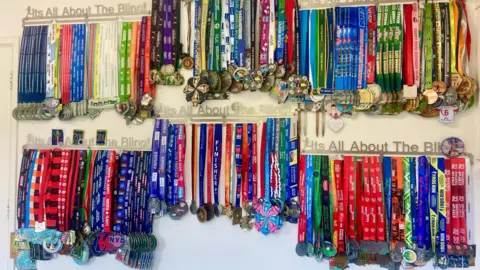 Elaine McAnulty A wall showing hundreds of medals hanging, they all have colourful lanyards and are arranged by date with markers highlighting significant achievements - 200, 300 etc... The rails they are hanging off say "It's all about the bling"