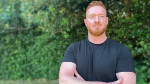 Ross Mackenzie stands with his arms crossed looking to the side with a serious expression