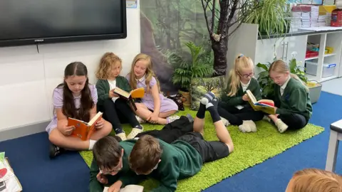 Henshaw C of E Primary School Seven children are reading. Two boys are lying on the floor, with a single girl to their right. Behind them are two pairs of girls sharing a book. They are on a bright green carpet and sitting at the base of a plastic tree.