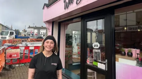 Girl with blonde hair wearing a black headband. She is standing outside of a cafe which has pink painting. Behind her is building works.