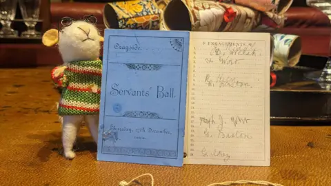 National Trust Small dainty card with one side blue and the other a ruled page with each row numbered. The blue side says "Servants' Ball" and the ruled page has people's names written against the numbers of different dances. Next to the card stands a little white felt mouse, with little metal-rimmed glasses and a green striped knitted jumper.