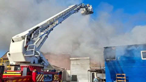 West Midlands Fire Service A white aerial platform with firefighters on it, they are spraying water down onto a fire, where you can see a significant amount of billowing smoke coming from a factory building